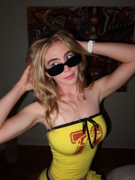 Grace Charis wearing black sunglasses and a yellow tube top posing with hands in her hair.
