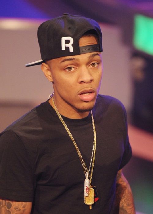 Bow Wow wearing a black t-shirt and black cap with a gold chain, looking at the camera.
