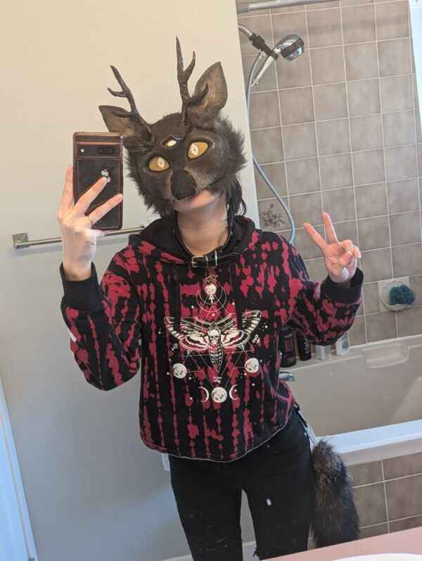 Person taking a mirror selfie while wearing a dark deer therian mask with antlers and glowing yellow eyes, dressed in a red and black hoodie in a bathroom setting.