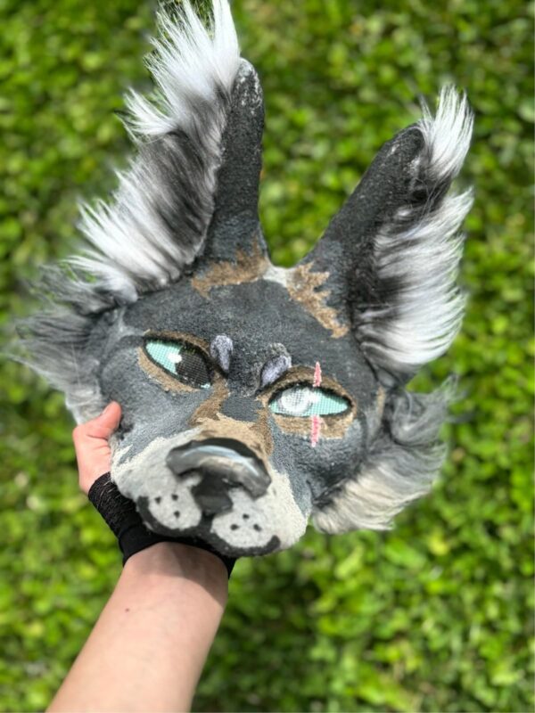 Handmade gray wolf therian mask with fluffy white and black fur, green mesh eyes, and detailed facial markings, held in a person’s hand outdoors against a green grass background.